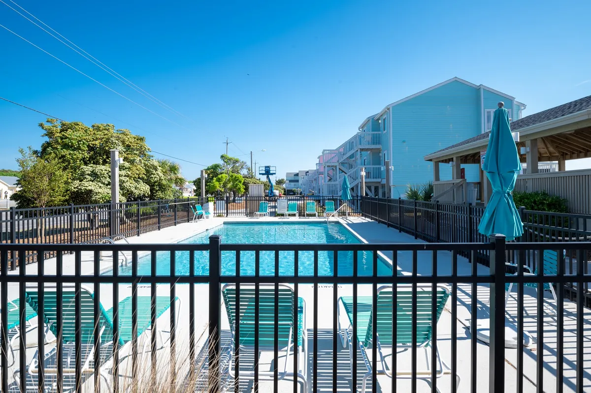 The image captures a sparkling swimming pool flanked by lounge chairs and shade umbrellas, all enclosed within a tidy fence. The scene suggests opportunities for swimming, sunbathing, and relaxed socializing in a bright, outdoor setting.