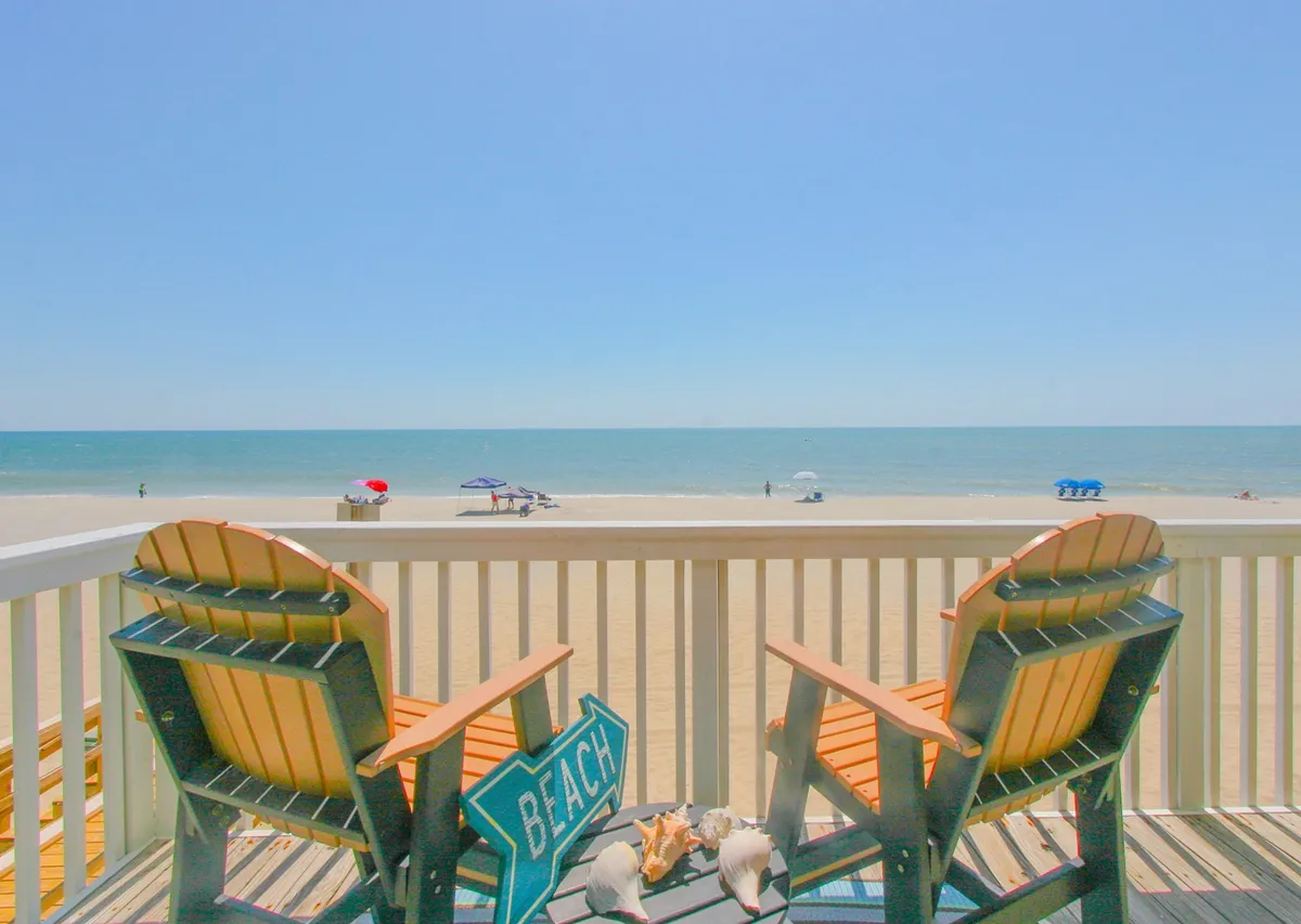 A serene beachfront scene with adirondack chairs set in soft sand, a beach sign pointing the way, ocean waves in the distance, and colorful umbrellas offering shade. The image captures the inviting expanse of shoreline perfect for families to relax, build sandcastles, and enjoy seaside activities.
