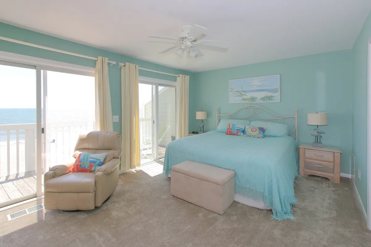 This image captures the primary king suite featuring a king-sized bed with turquoise bedding, expansive windows showcasing an ocean view, and a comfortable armchair positioned for relaxation or reading. The tranquil light and scenic backdrop evoke a sense of privacy and restful retreat.