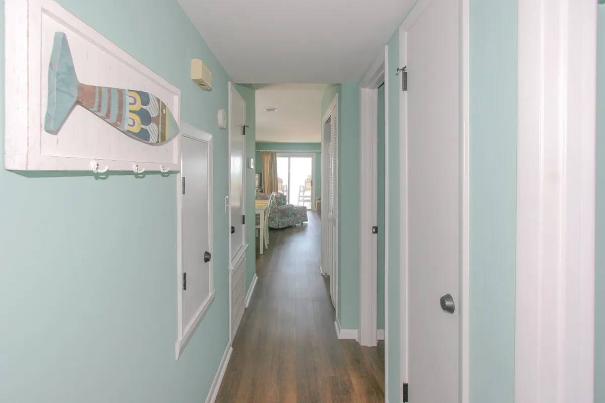 This image features a light-filled entryway with colorful fish wall art, light wood flooring, and an open door that leads into the living area. The space feels welcoming and organized, with clear pathways and a relaxed coastal vibe.
