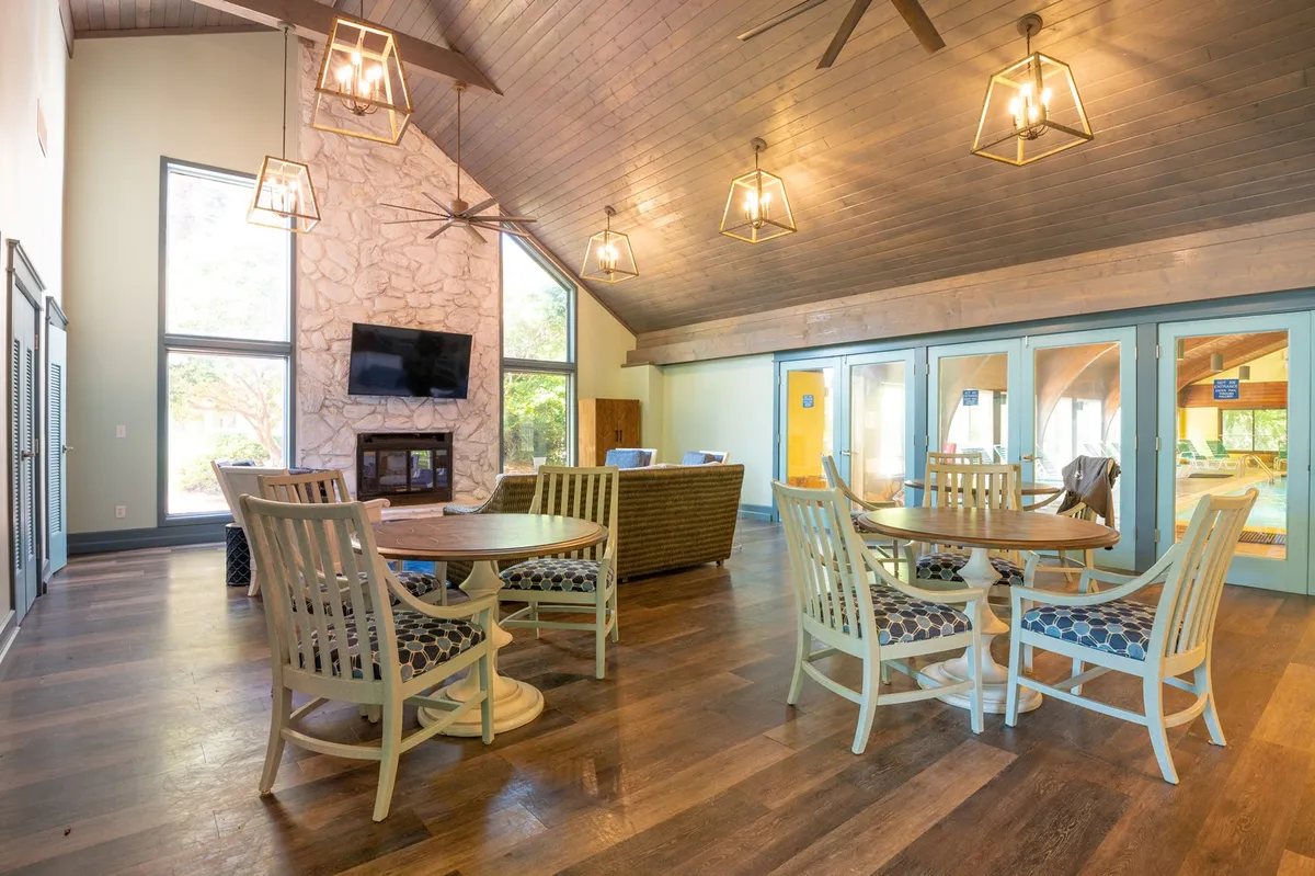 A spacious, communal recreation center interior with large windows, a cozy stone fireplace, circular tables for gatherings, and relaxed seating. The setting is perfect for socializing, playing games, or unwinding after a day of adventure.