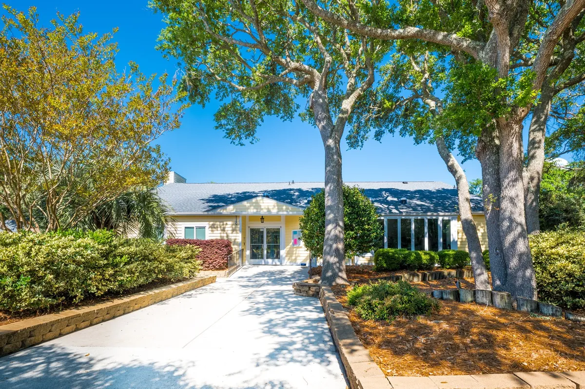 A clear view of the property's front entrance, framed by large trees and a landscaped garden. The designated parking area is subtly visible, hinting at both assigned and guest parking options. The entry looks secure and welcoming.