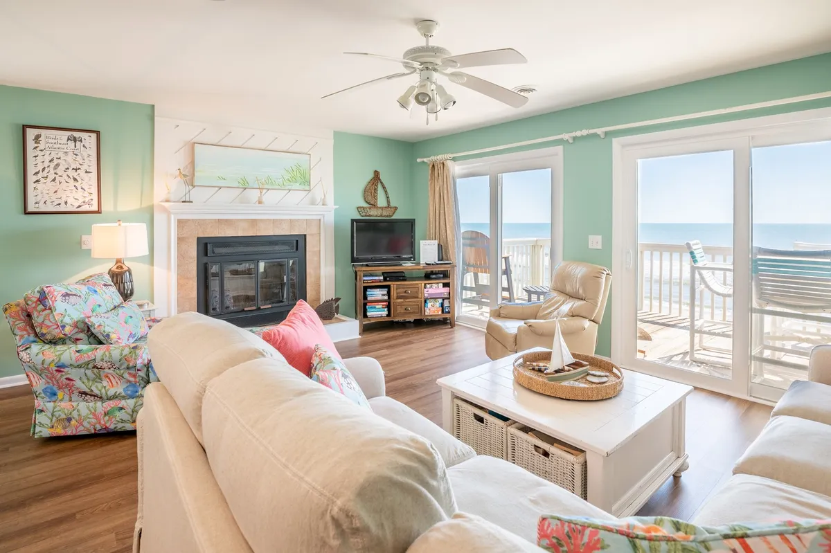The image shows a beautifully decorated living room in a Kure Beach oceanfront condo. Large windows frame an unobstructed view of the Atlantic, flooding the space with natural light. Comfortable seating is arranged to face both the cozy fireplace and the ocean, inviting relaxation. Beach-themed artwork and tasteful coastal decor add a fresh, inviting vibe. The space is perfect for gathering, unwinding with a book, enjoying breathtaking sunrises, or sharing sunsets with loved ones.