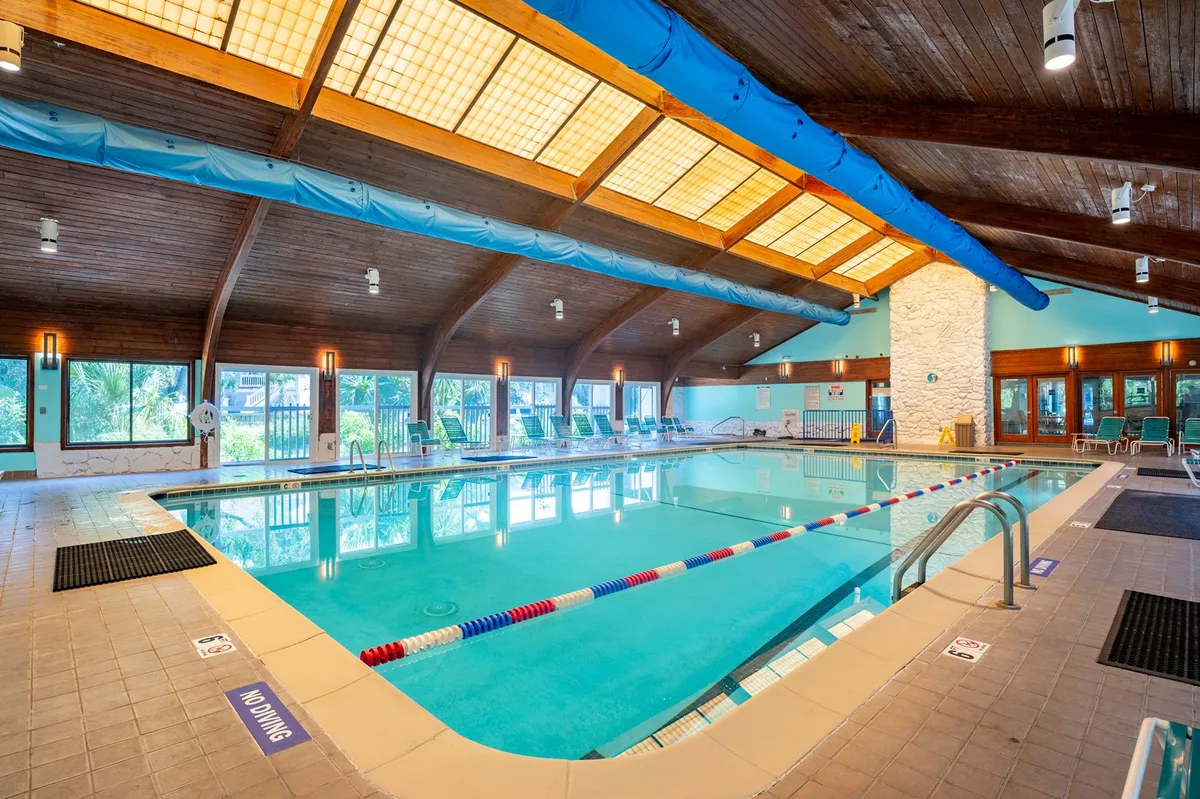 A modern indoor swimming pool with wooden beams overhead, large windows that let in natural light, and comfortable lounge chairs lining the edge. The ambiance is serene and well-suited for year-round enjoyment.