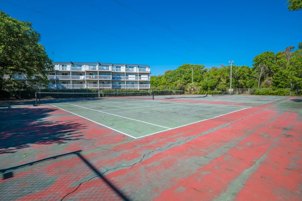 This image displays a vibrant tennis court surrounded by lush greenery, with a nearby apartment building visible. The tennis court is well-maintained and bathed in sunlight, offering an inviting space for active play and socializing outdoors.