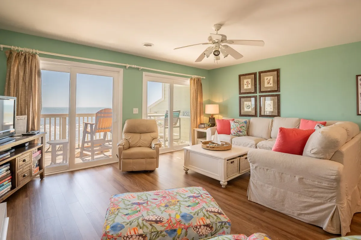 A spacious living room featuring a colorful couch with vibrant pillows, floral ottoman, large wooden coffee table, and expansive windows that frame an inviting ocean view. The décor and space evoke a cheerful, beachy, and welcoming vibe, ideal for lounging or socializing.