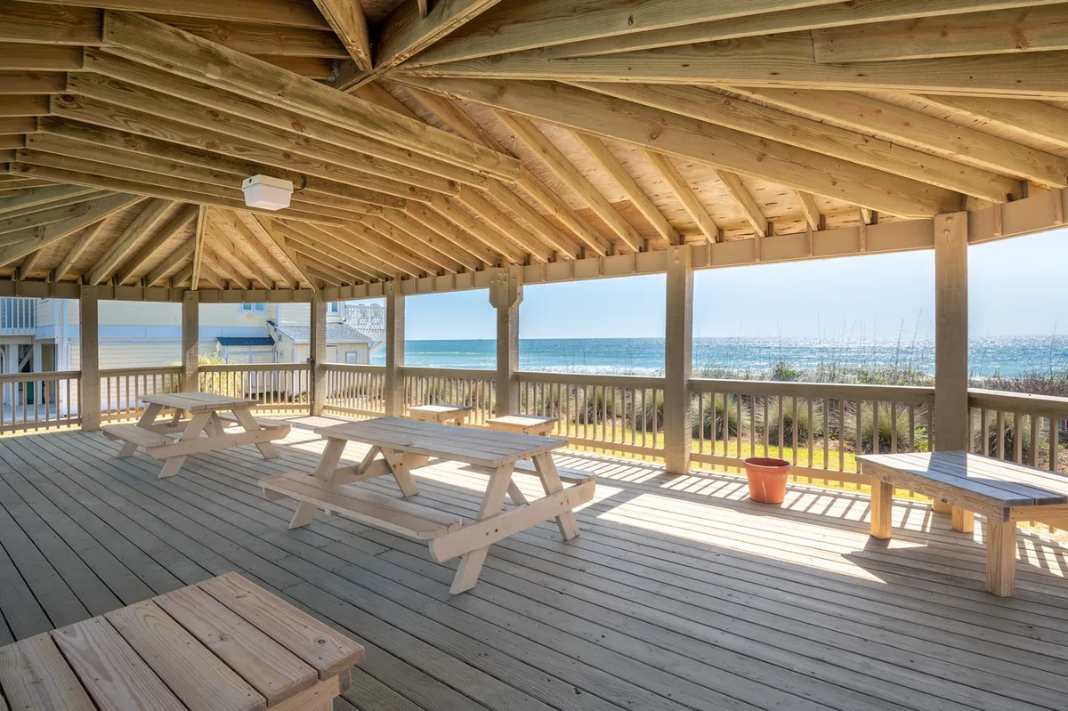 A wide wooden deck with picnic tables overlooks the ocean, offering expansive views of the water. The setting is open and inviting, perfect for group meals, sunset watching, and family gatherings. The deck is surrounded by natural greenery, emphasizing its tranquil seaside atmosphere.